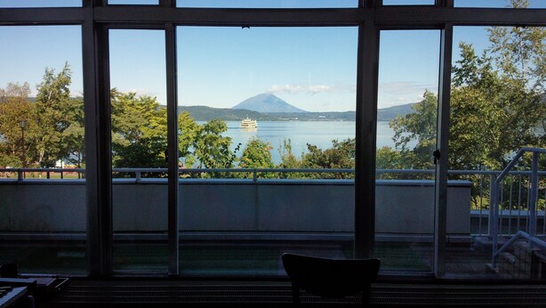 A view from a balcony with a chair, overlooking a lake with a boat and a mountain in the distance. - Granvillage Toya Daiwa Ryokan Annex (Toyako)