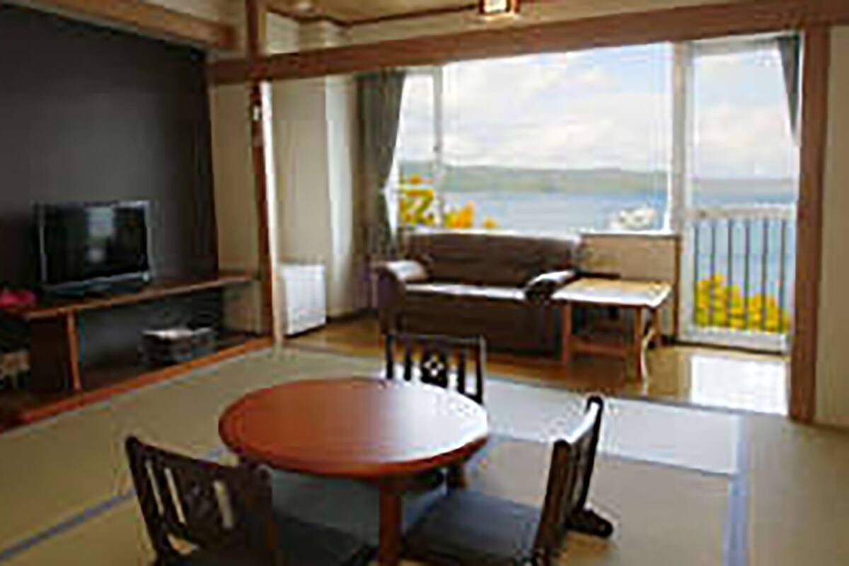 Un salon avec une télévision, un canapé, une table et des chaises. Il y a un balcon avec vue sur la mer.