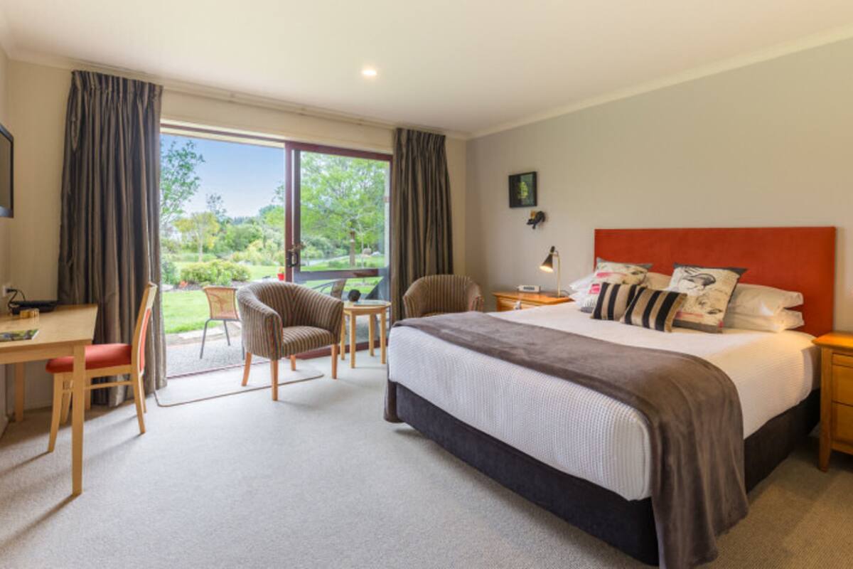 Ein Hotelzimmer mit einem großen Bett, einem Schreibtisch und einem Stuhl. Ein Fenster mit Vorhängen bietet Blick auf Grünflächen.