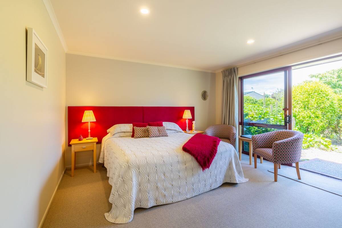 Ein Hotelzimmer mit einem großen Bett, zwei Nachttischen mit Lampen, einem Sessel und Blick auf Grün durch ein großes Fenster.