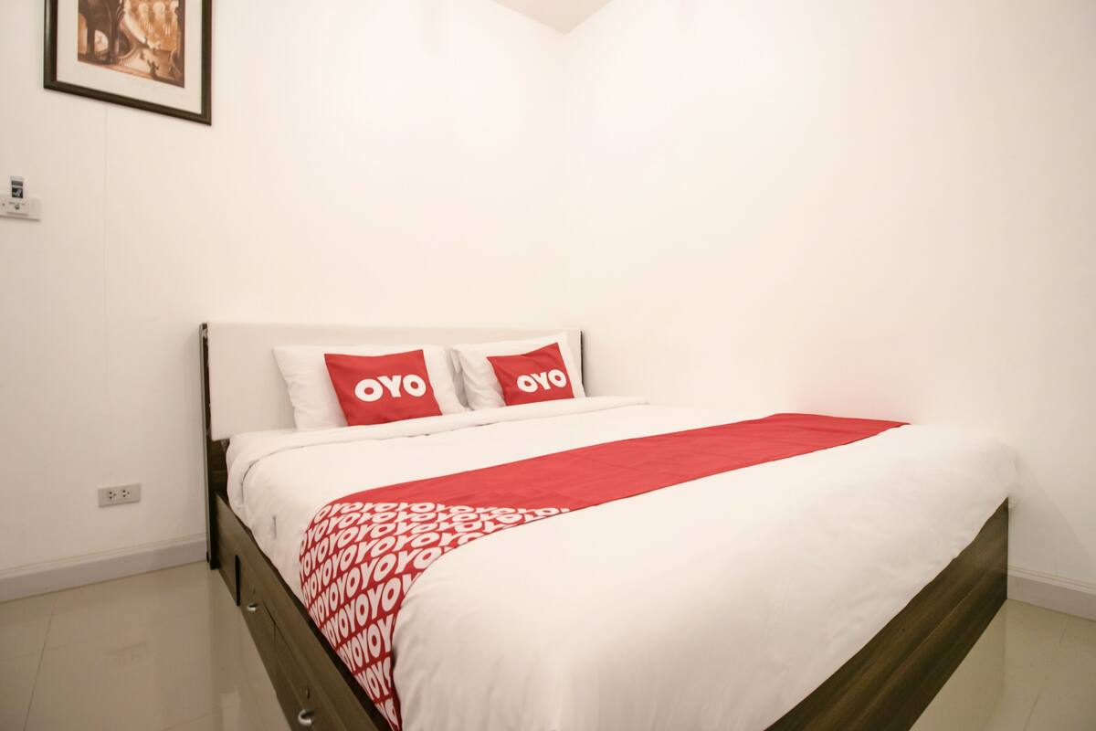 A neatly made bed with a white and red bedspread and pillows, a wooden headboard, and a white wall with a framed picture.