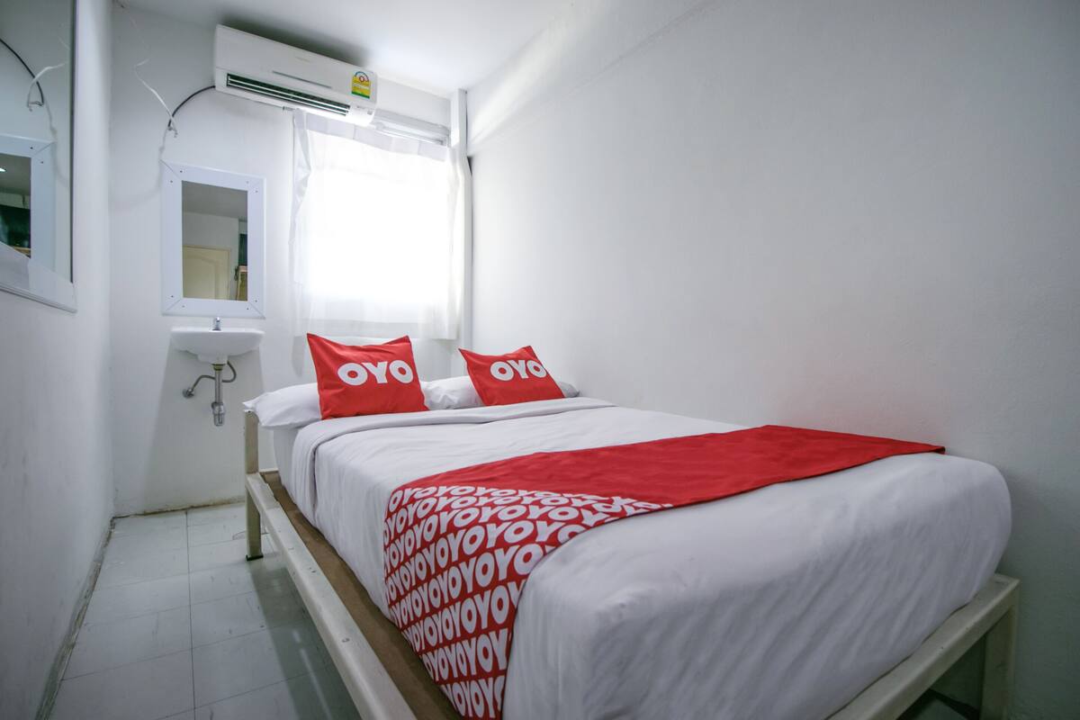 A single-bed room with a white bedspread, two red pillows, a white bedside table, and a white sink.