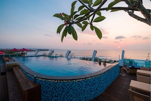 Une piscine à débordement avec une bordure carrelée, une terrasse en bois et des chaises longues.