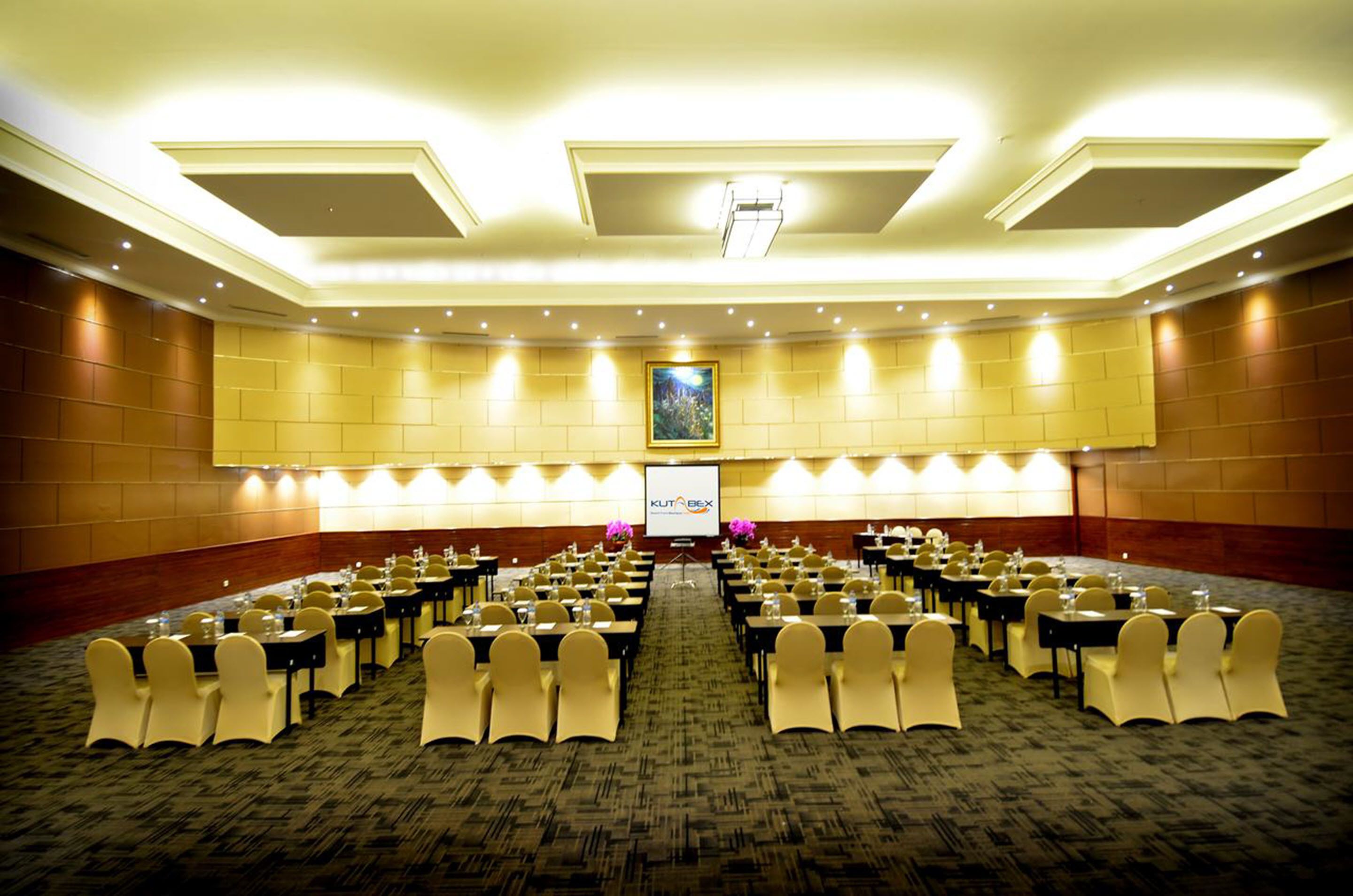 A conference room with rows of tables and chairs, a projector screen, and a large framed picture on the wall.
