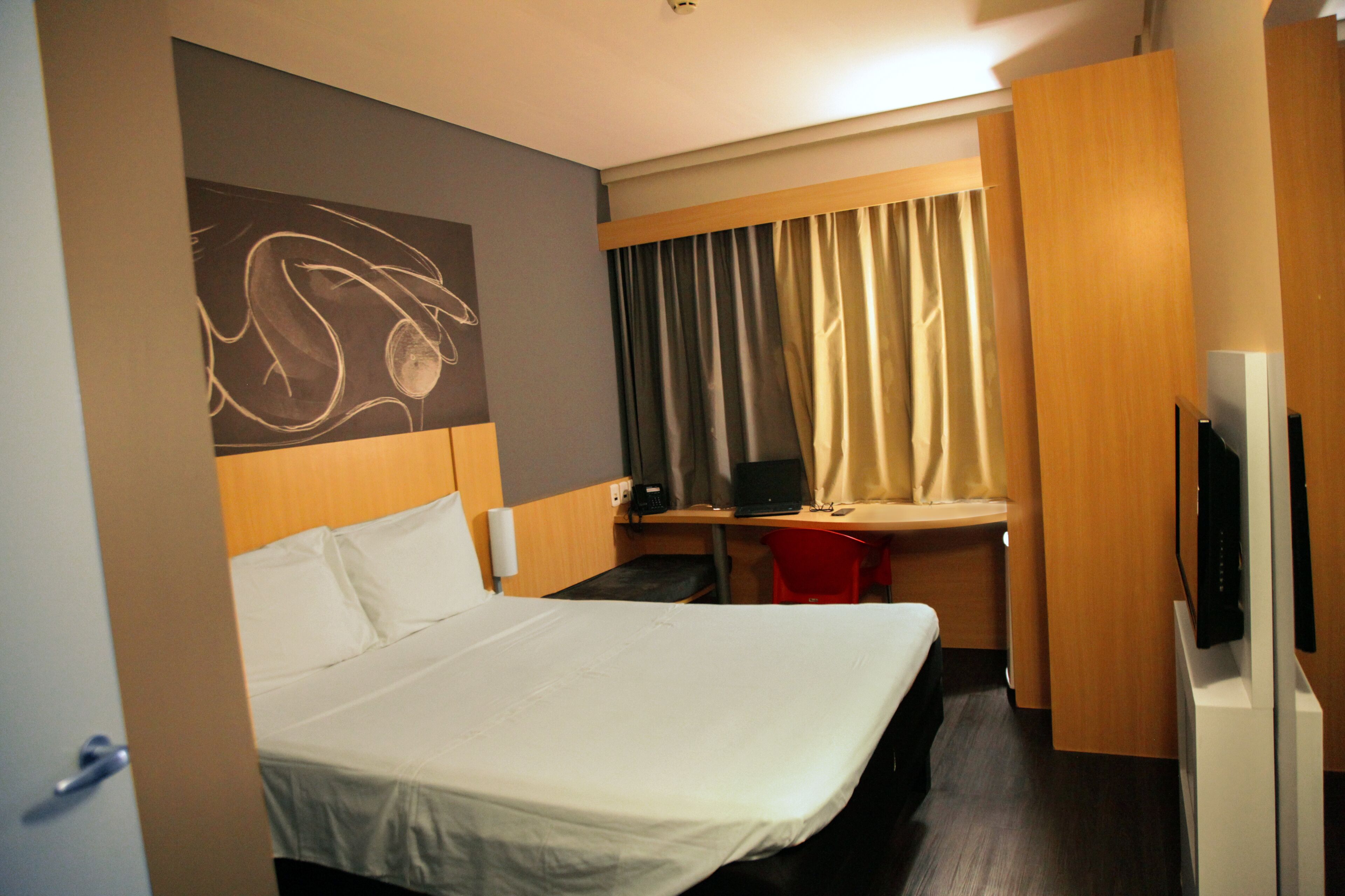 A hotel room with a bed, a desk with a laptop, a TV, and a red chair.