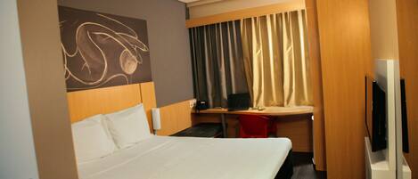 A hotel room with a bed, a desk with a laptop, a TV, and a red chair.