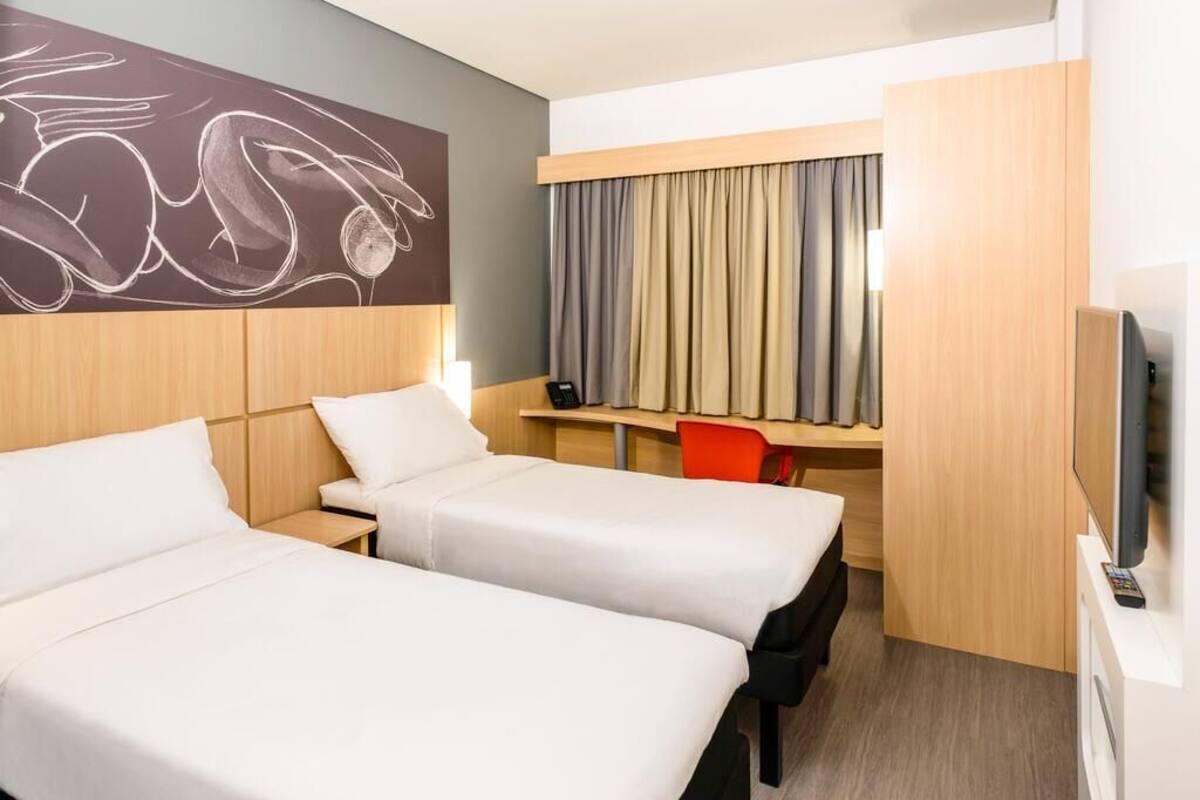A hotel room with two beds, a desk, a chair, a TV, and a window with curtains.