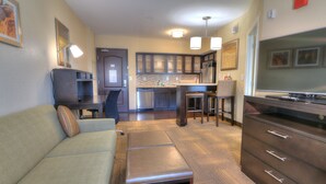 A modern living room with a sofa, a coffee table, and a TV. There is a kitchen area with cabinets and a refrigerator in the background. - Staybridge Suites Knoxville-West by IHG (Farragut)