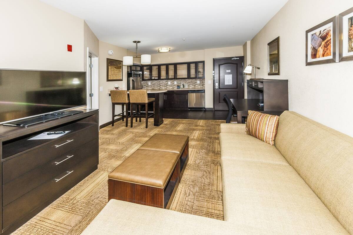 A modern living room with a large flat-screen TV, a sofa, a coffee table, and a dining area with chairs and a kitchen in the background.