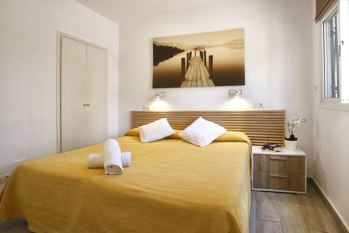A neatly made bed with a yellow bedspread, two white pillows, and a rolled towel. There is a wooden headboard, a bedside table with a lamp, and a wall-mounted painting of a dock.