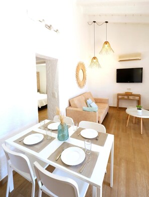A modern dining area with a white table set for four, a sofa, a TV, and a small side table with a plant. - Naika Studios & Apartments (Calvia)