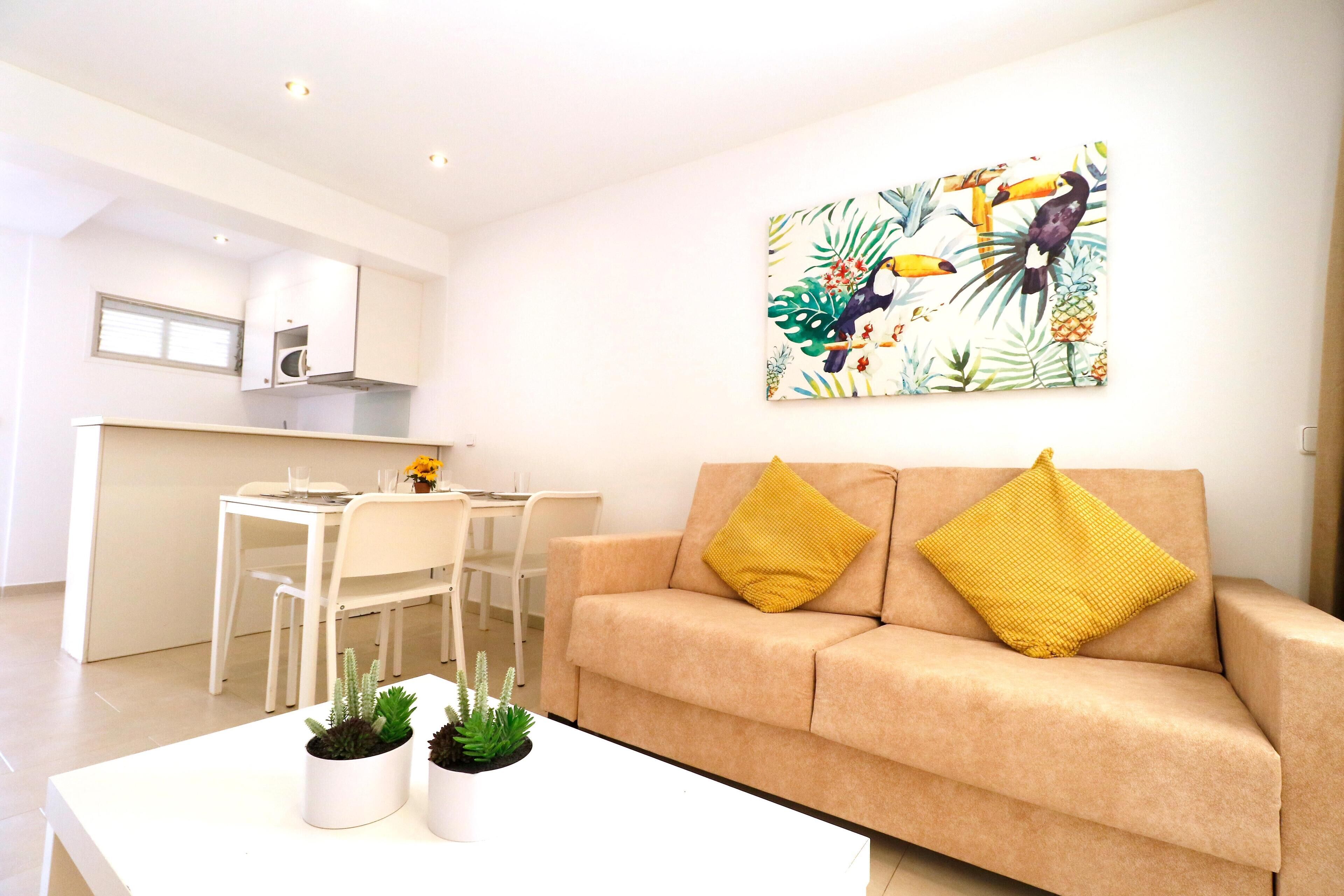 A modern living room with a beige sofa, two yellow cushions, a white coffee table with potted plants, a dining area with a white table and chairs, and a kitchen with white cabinets and a painting of tropical birds.