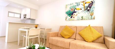 A modern living room with a beige sofa, two yellow cushions, a white coffee table with potted plants, a dining area with a white table and chairs, and a kitchen with white cabinets and a painting of tropical birds.