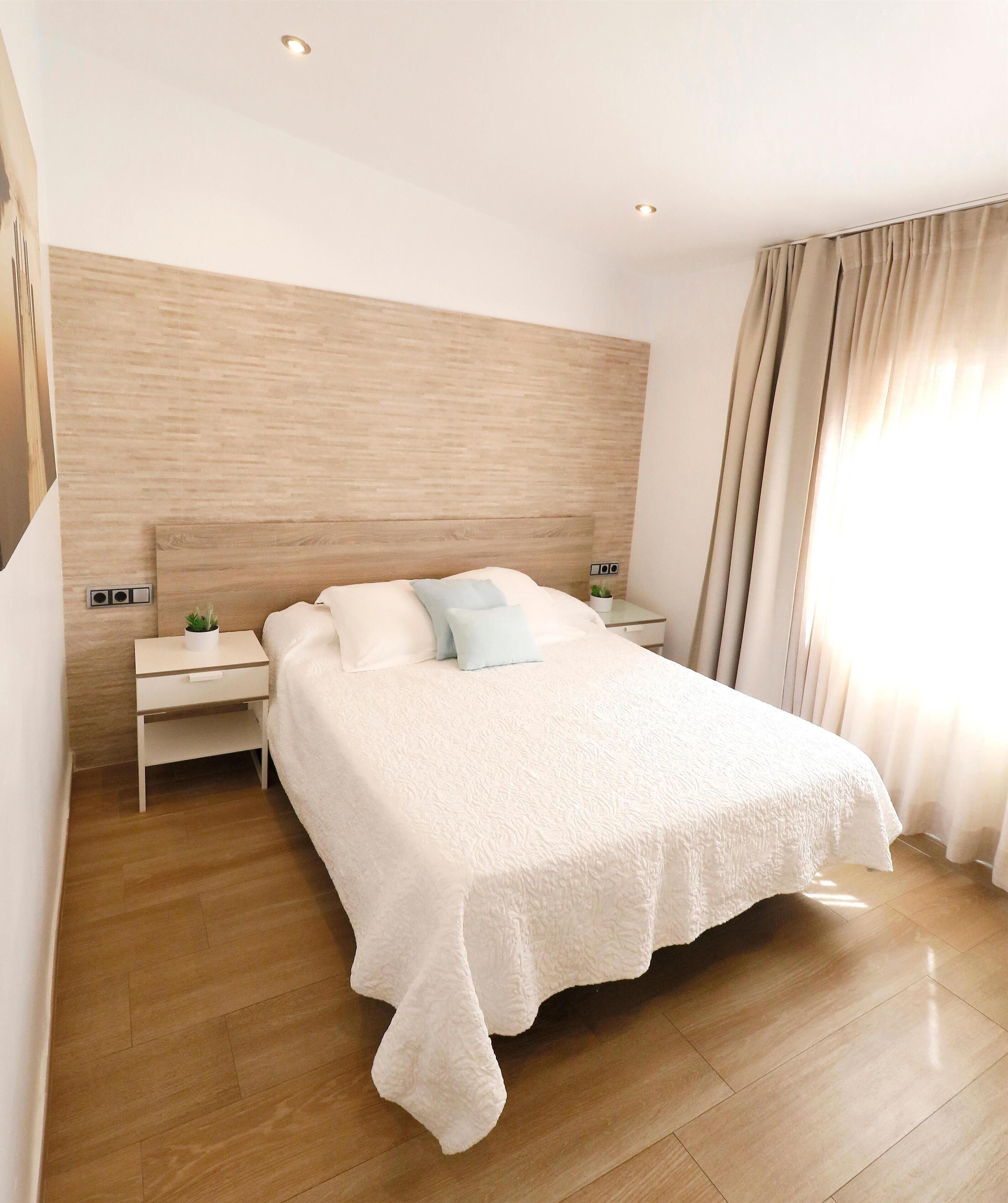 Un dormitorio moderno con una cama grande, cabecera de madera, mesitas de noche con lámparas y vistas al exterior a través de cortinas.