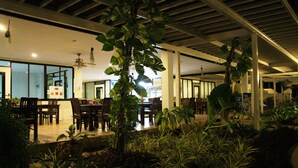 An indoor dining area with tables and chairs, surrounded by potted plants and a ceiling fan.