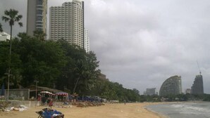 A beach with a sandy shore, palm trees, and a few beach chairs. In the background, there are tall buildings and a cloudy sky.