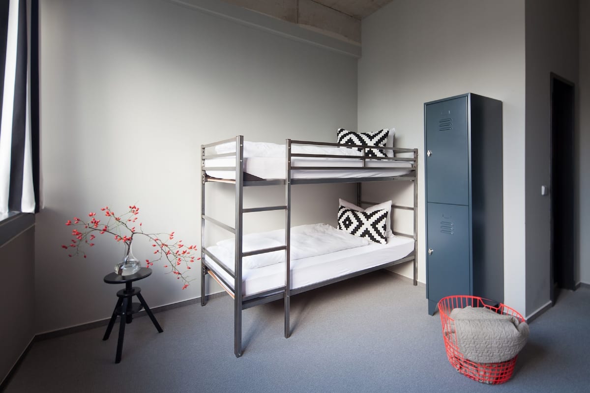 Ein Etagenbett mit zwei Kissen, ein kleiner Tisch mit einer Vase, ein roter Korb und ein grauer Schrank in einem Zimmer mit schlichter Wand und Teppich.