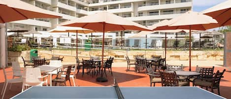 An outdoor table tennis table with surrounding chairs and umbrellas, set against a backdrop of a multi-story building with balconies.