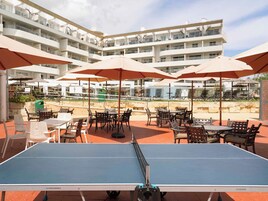 Una mesa de tenis de exterior con sillas y sombrillas alrededor, frente a un edificio de varios pisos con balcones.