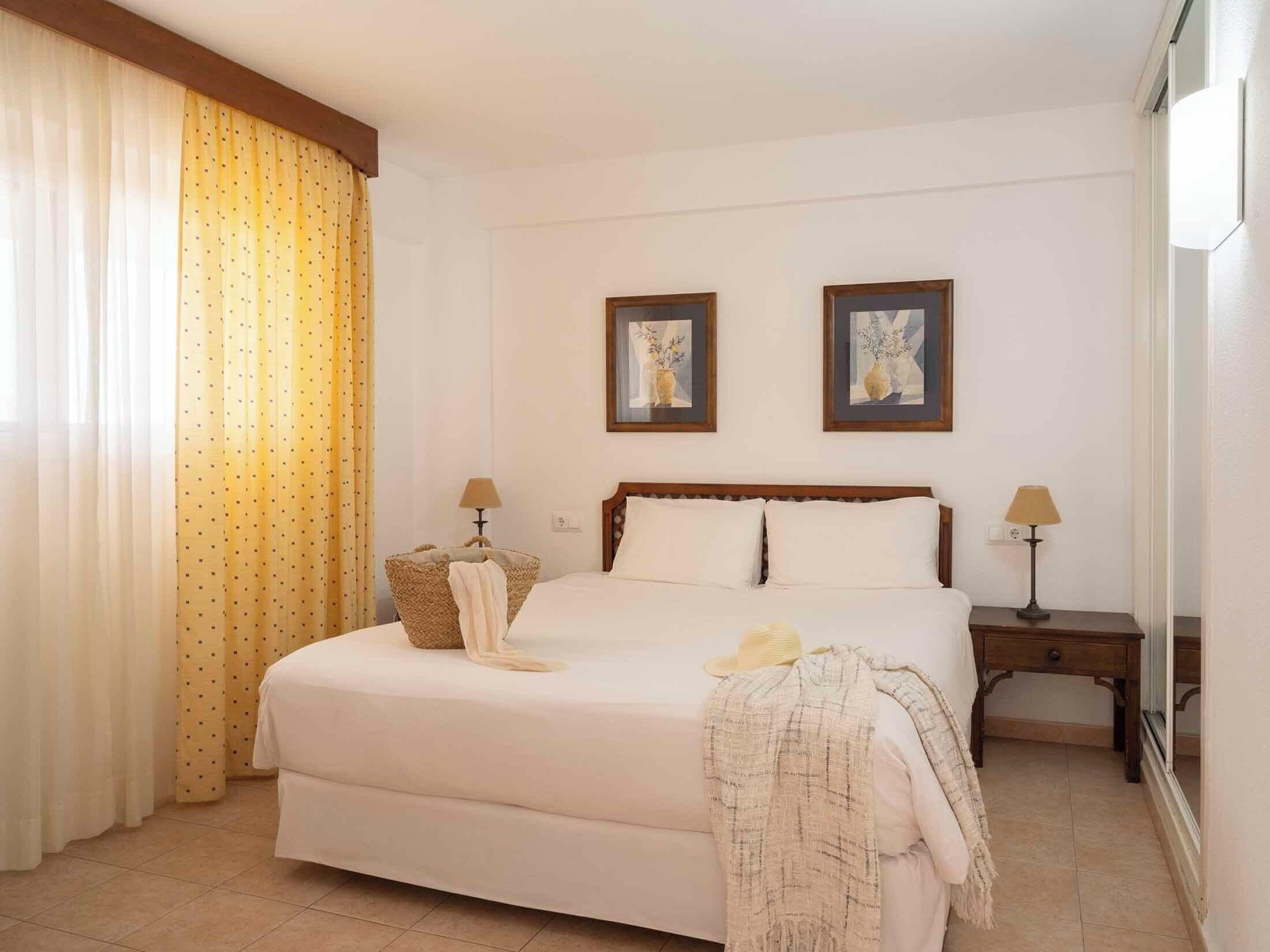 A hotel room with a bed, two bedside tables, a lamp on each table, a large window with curtains, and two framed pictures on the wall.