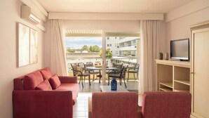 Habitación de hotel con sofá rojo, televisor sobre un mueble bajo, balcón con mobiliario exterior y vistas a un edificio y árboles.