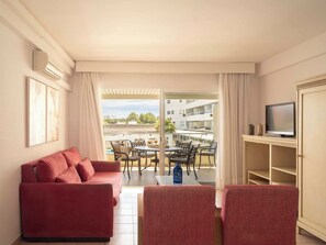 Habitación de hotel con sofá rojo, televisor sobre un mueble bajo, balcón con mobiliario exterior y vistas a un edificio y árboles.