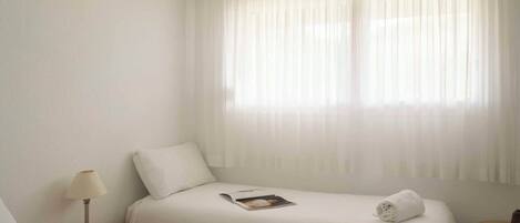 A hotel room with two beds, a nightstand, a lamp, and a window with sheer curtains.