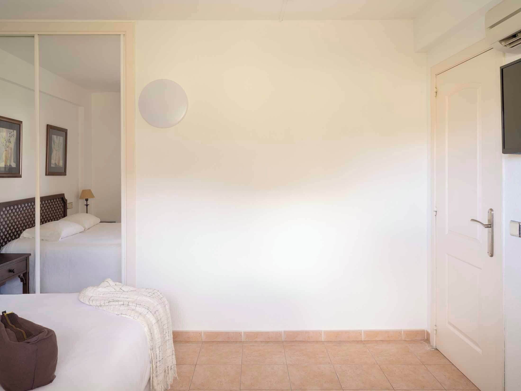 Habitación de hotel con cama, mesita de noche, lámpara, televisor montado en la pared y una puerta.