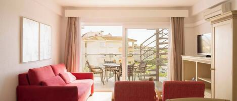 Habitación de hotel con un sofá rojo, una mesa de comedor con sillas, un televisor y vistas a edificios a través de un ventanal.