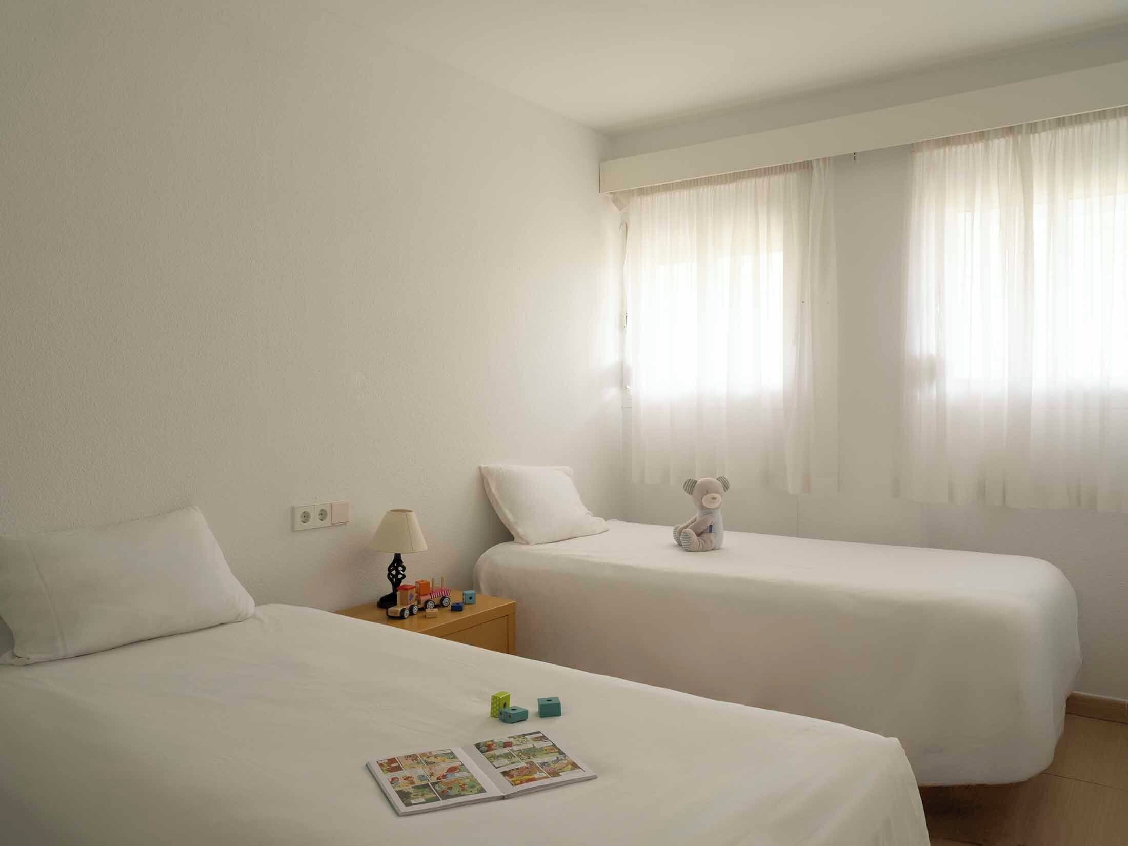 Habitación de hotel con dos camas, una mesita de noche con una lámpara, un osito de peluche y una revista en el suelo.