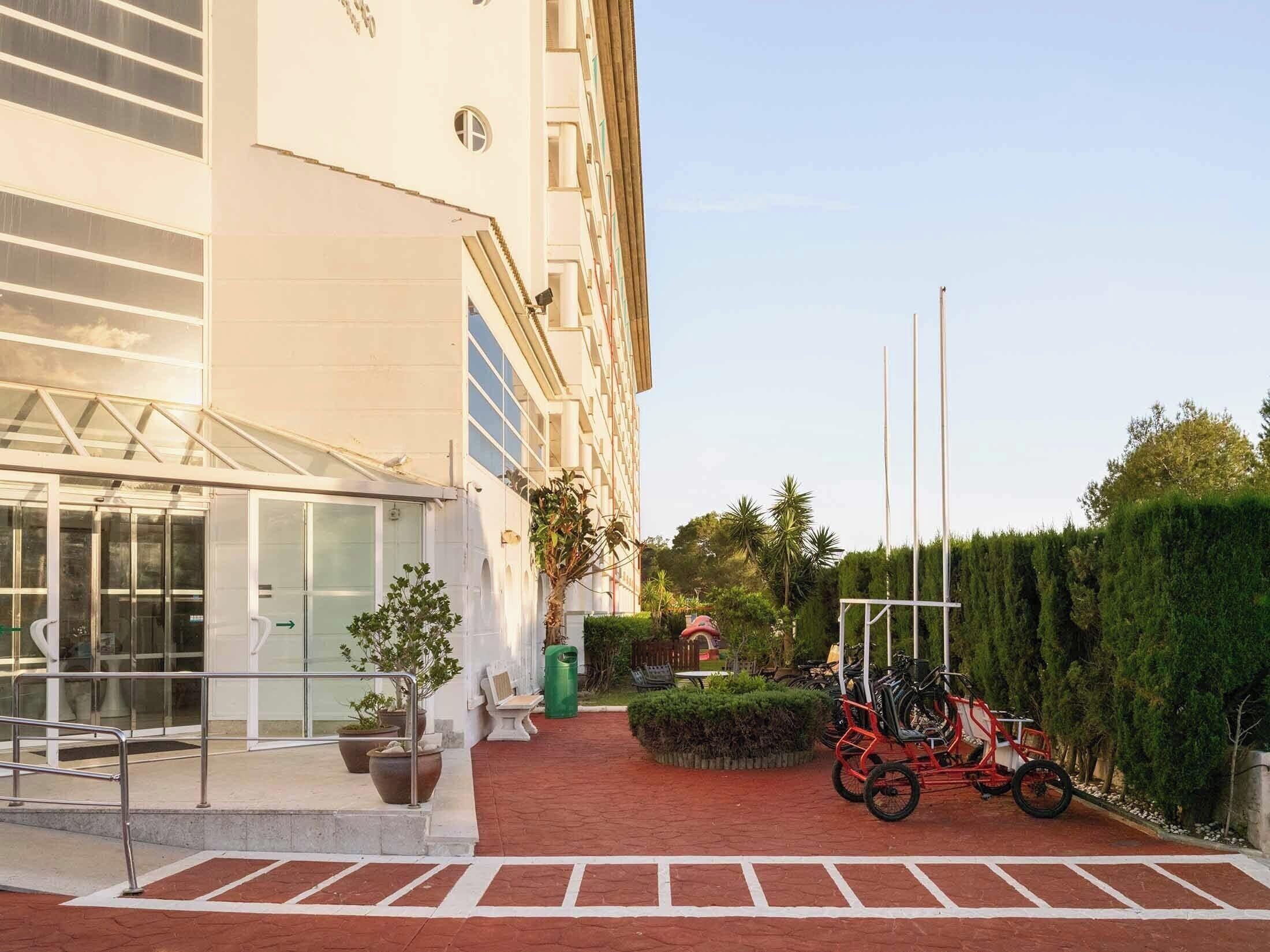 Entrada de hotel con una galería techada, plantas en macetas y un aparcamiento para bicicletas.