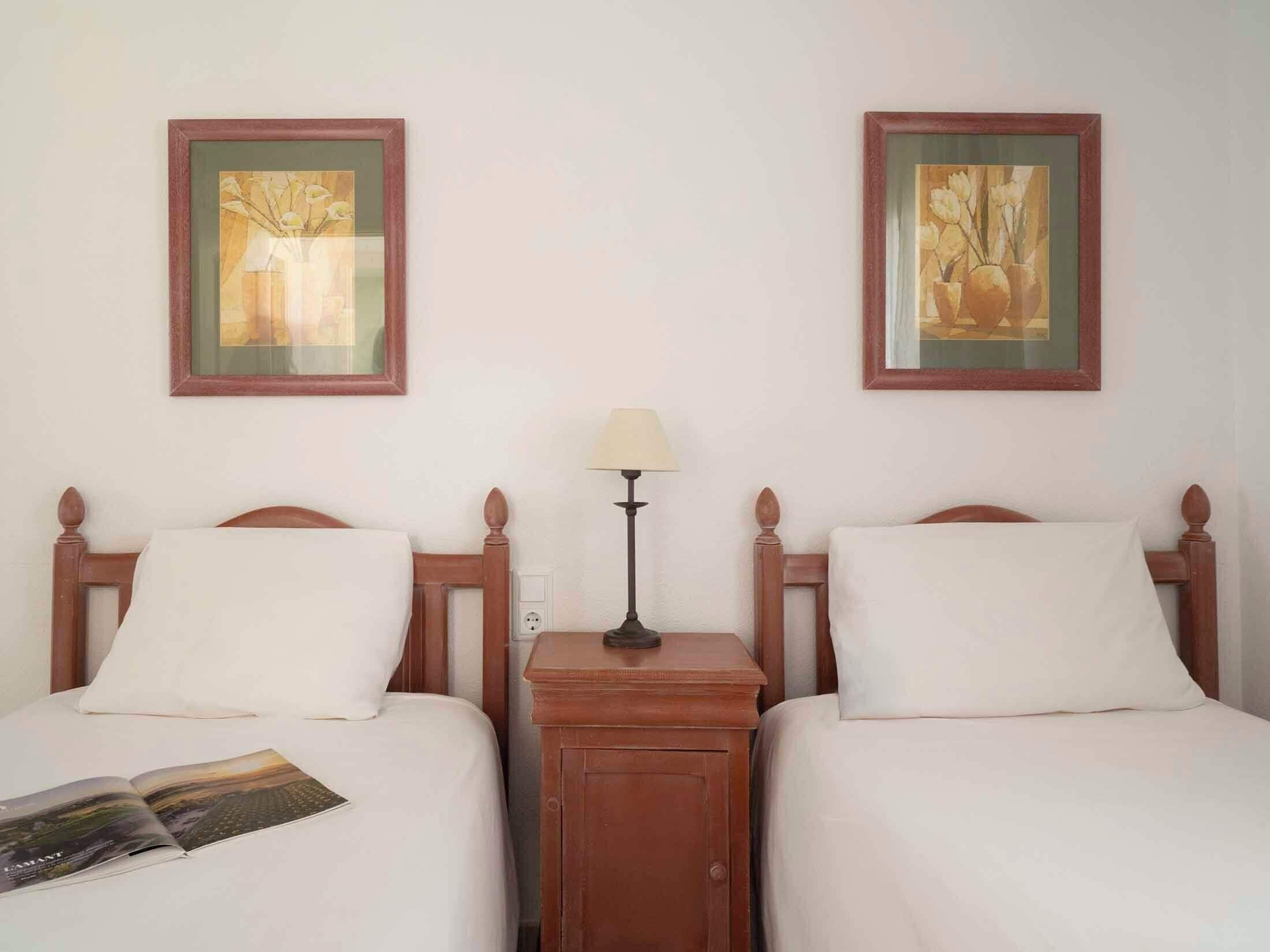 Una habitación de hotel con dos camas, una mesita de noche, una lámpara y dos cuadros en la pared.