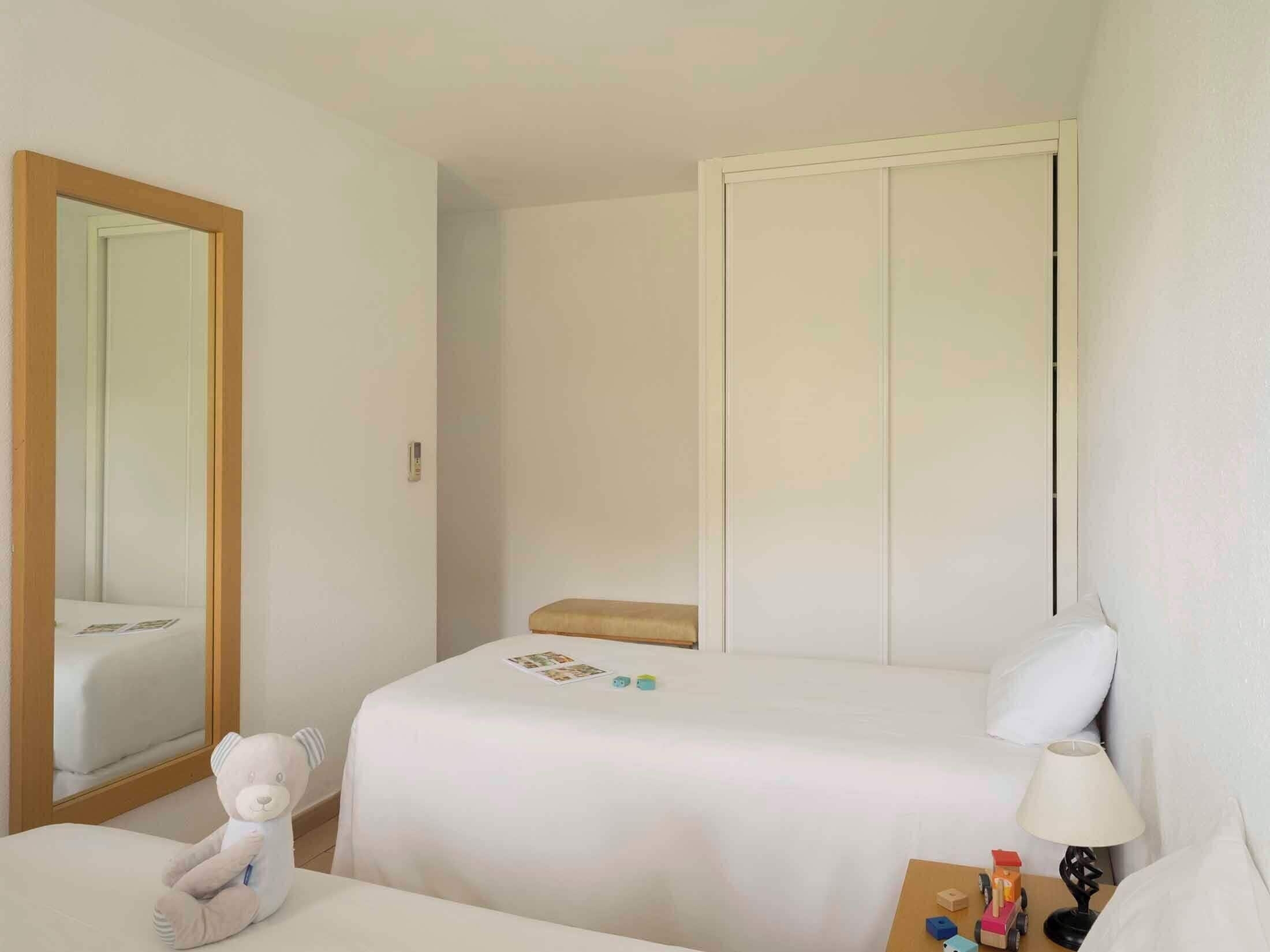 A hotel room with a bed, a bedside table with a lamp, a chair, a teddy bear, and a wardrobe.