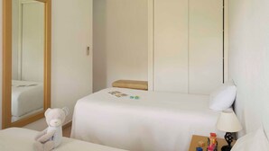 Una habitación de hotel con cama, una mesita de noche con lámpara, una silla, un osito de peluche y un armario.