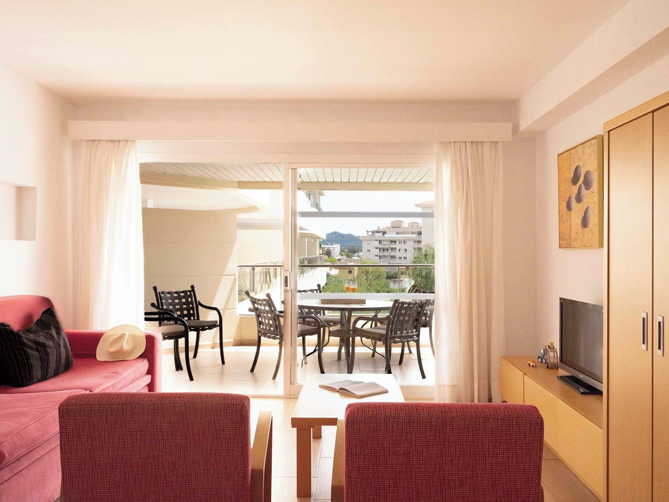Une pièce avec un canapé rouge, un téléviseur, une table à manger avec des chaises et un balcon donnant sur des immeubles.