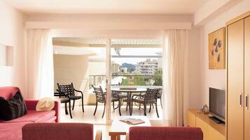 Une pièce avec un canapé rouge, un téléviseur, une table à manger avec des chaises et un balcon donnant sur des immeubles.
