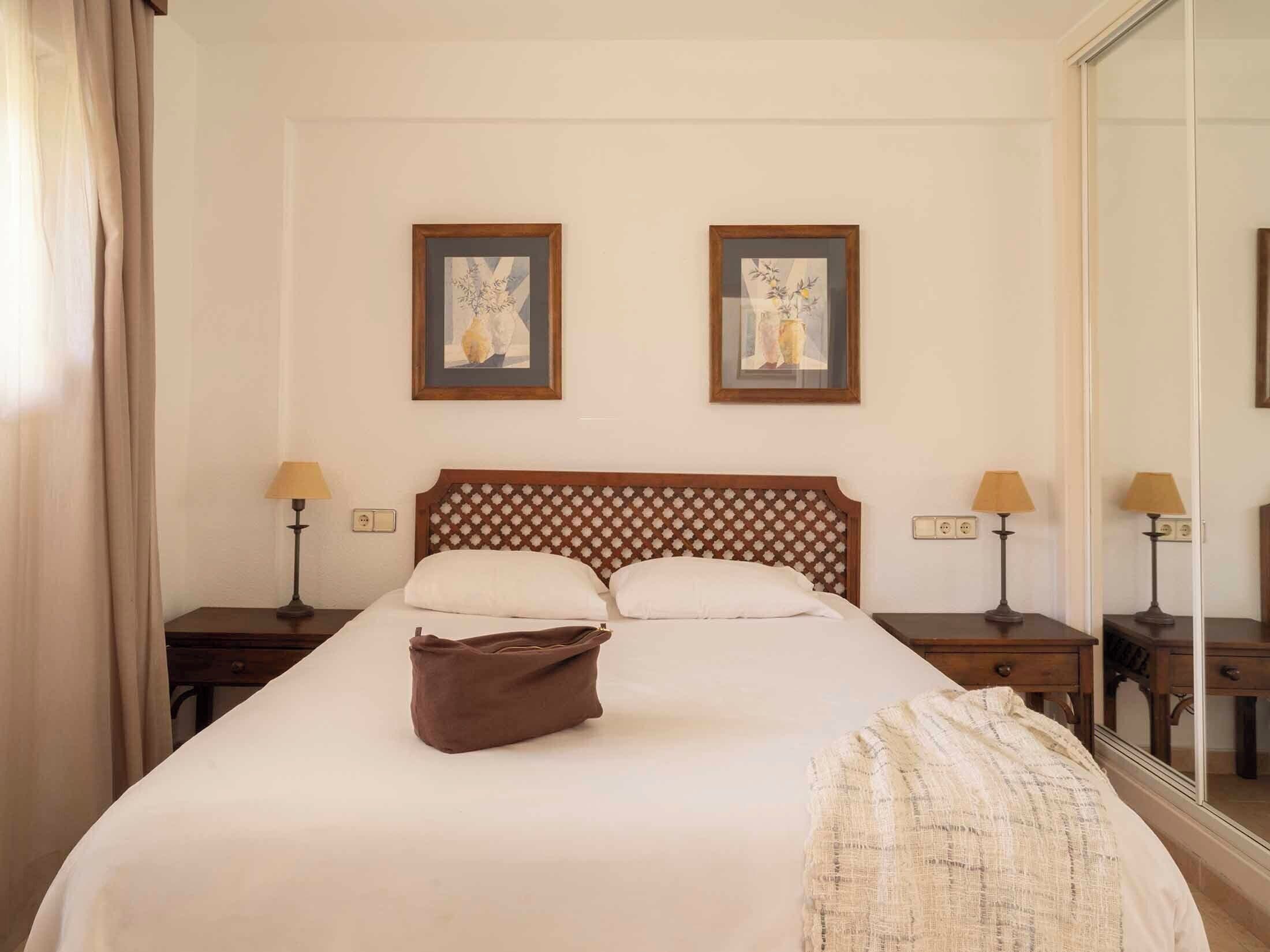 A hotel room with a large bed, two bedside tables with lamps, a wooden headboard, and two framed artworks on the wall.