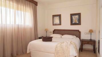 Una habitación de hotel con una cama, dos mesitas de noche con lámparas, un ventanal con cortinas y dos cuadros en la pared.