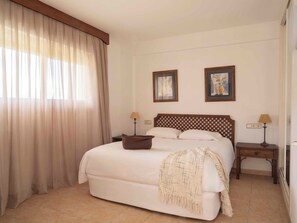 Una habitación de hotel con una cama, dos mesitas de noche con lámparas, un ventanal con cortinas y dos cuadros en la pared.