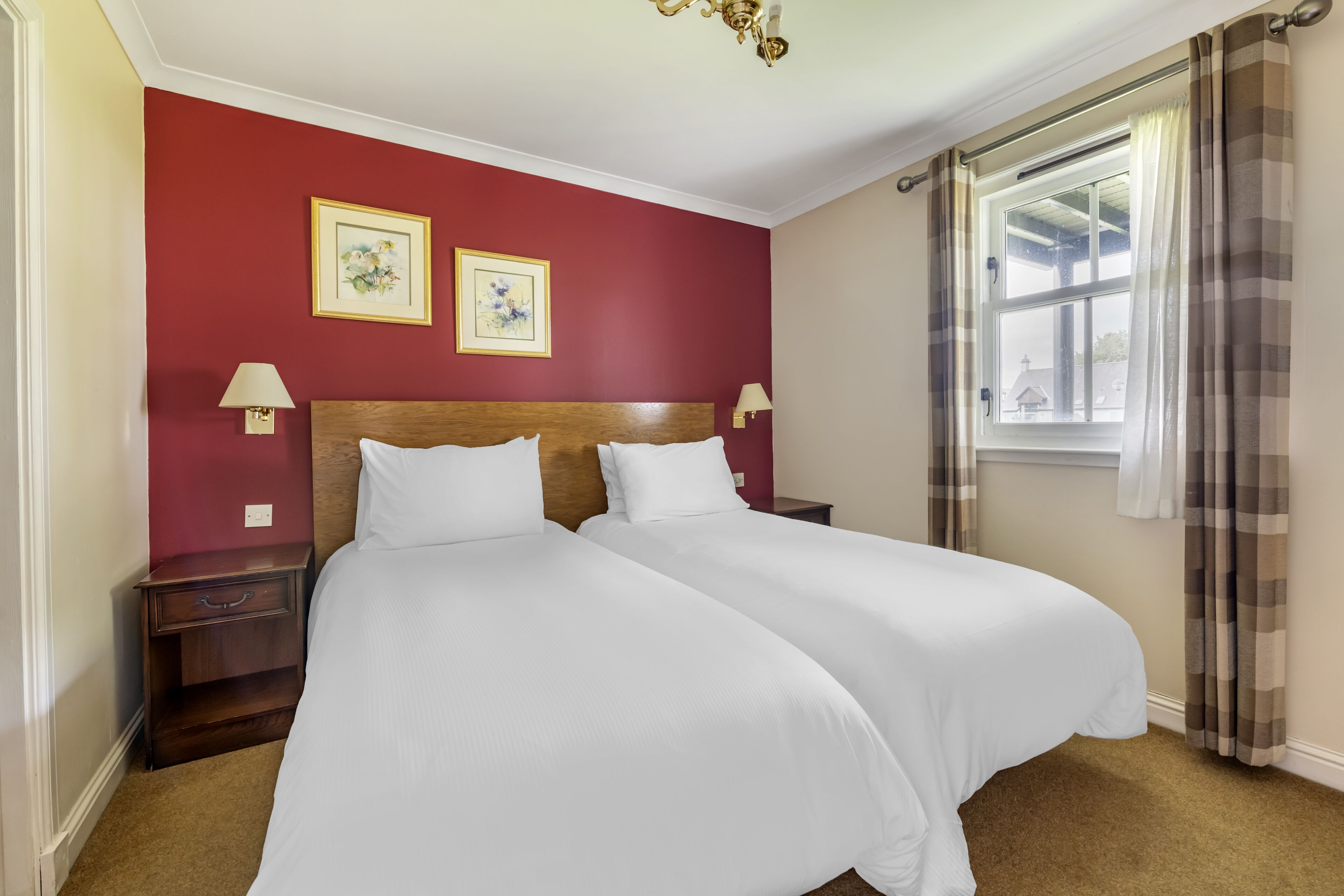 A hotel room with two beds, a red accent wall, two framed pictures, bedside lamps, a window with curtains, and a chandelier.