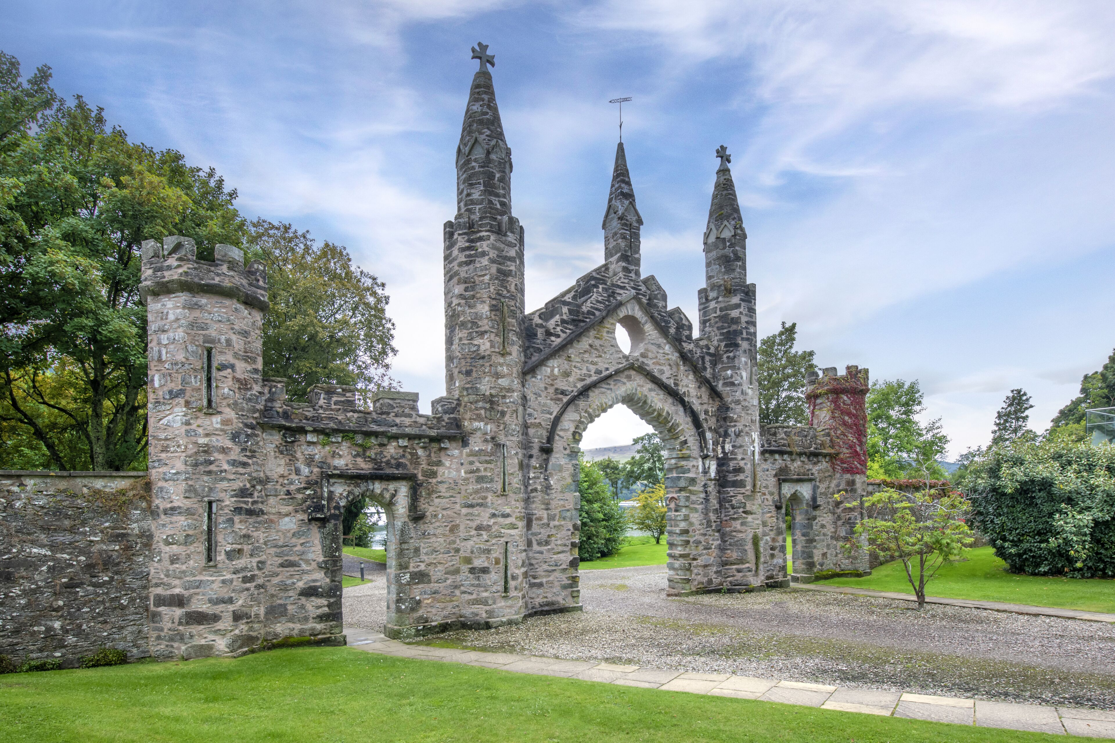 A historic stone building with a central archway and multiple spires, surrounded by a well-maintained lawn and trees.