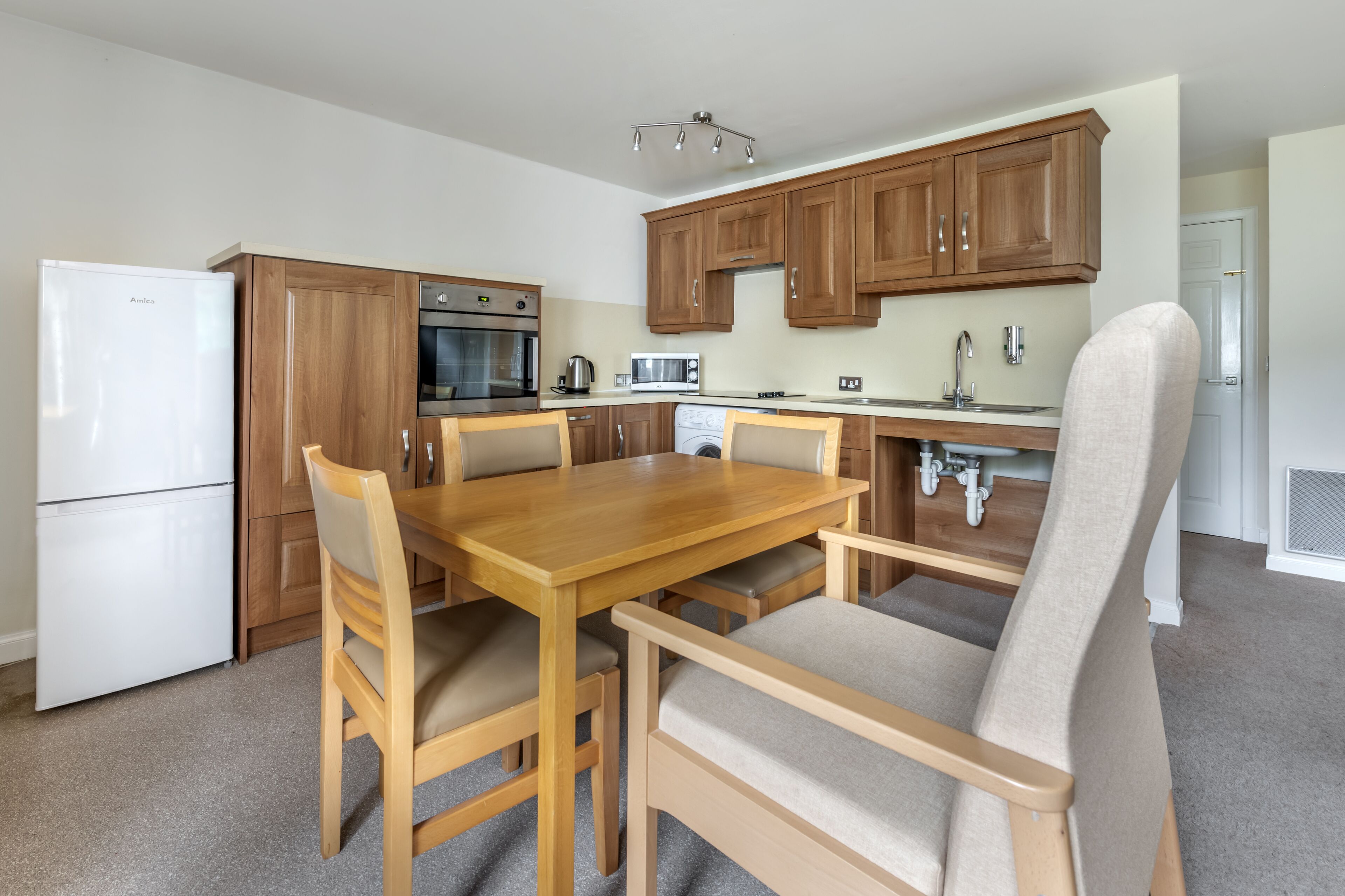 Una cocina moderna con armarios de madera, horno empotrado, microondas y un lavabo. Hay una mesa de comedor con sillas y un sillón grande con asiento acolchado.