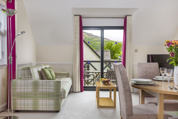 A room with a green plaid sofa, a dining table with chairs, a small wooden table with a bowl of fruit, and a view of a hillside through a glass door. - The Kenmore Club (Aberfeldy)