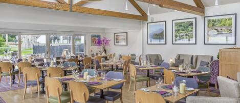 Un comedor con mesas y sillas de madera, cuadros en las paredes y grandes ventanales con vistas al exterior.