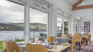 A dining area with tables set for guests, large windows offering a view of the water and hills, and a cozy interior with wooden beams and a plaid carpet. - The Kenmore Club (Aberfeldy)