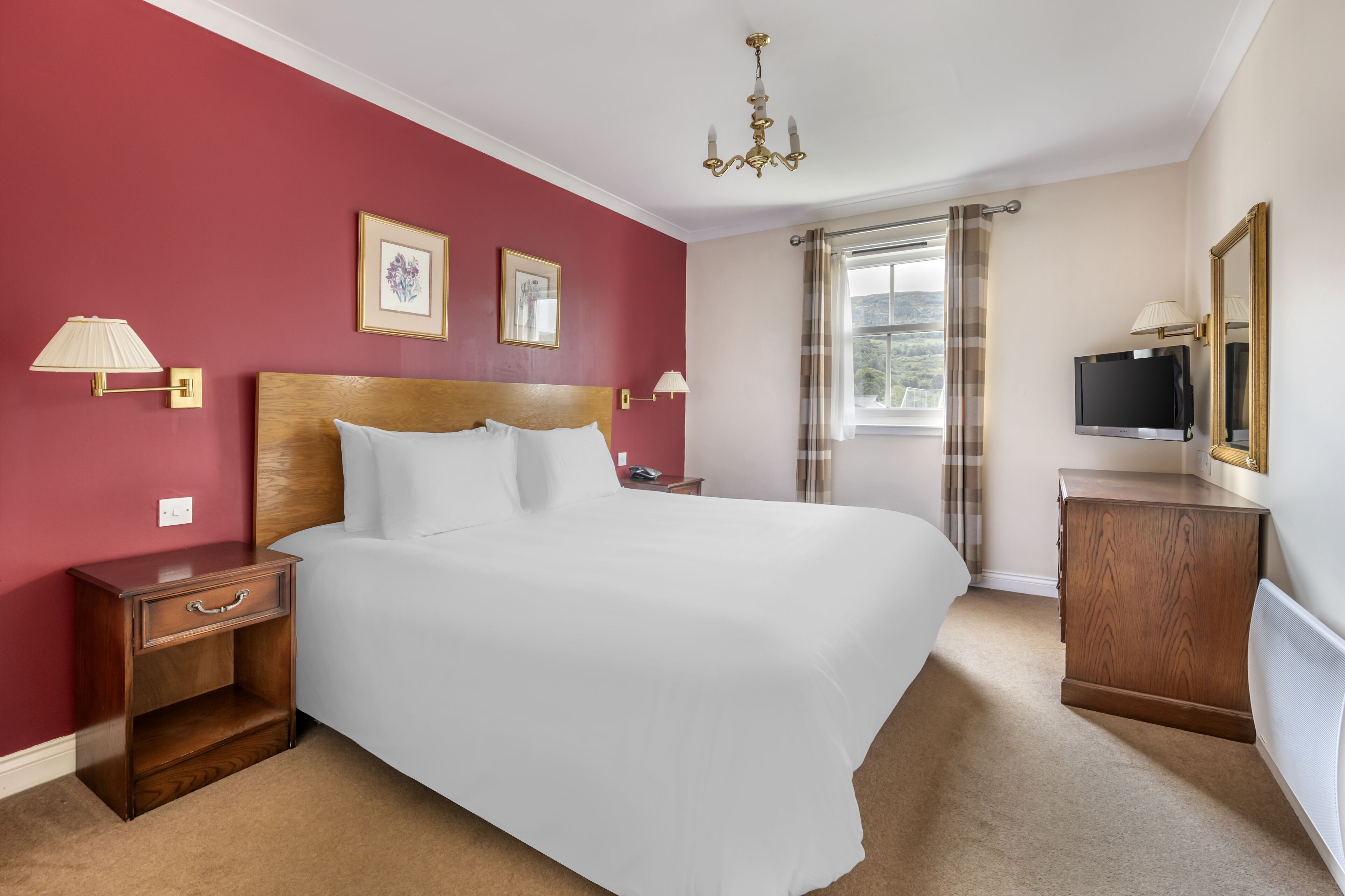 Una camera d'albergo con un letto grande, una testiera in legno, due comodini con lampade, una televisione, uno specchio e una finestra con tende.