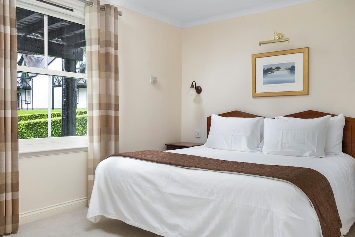 A hotel room with a bed, a window with curtains, a bedside table, a wall-mounted lamp, and a framed picture.