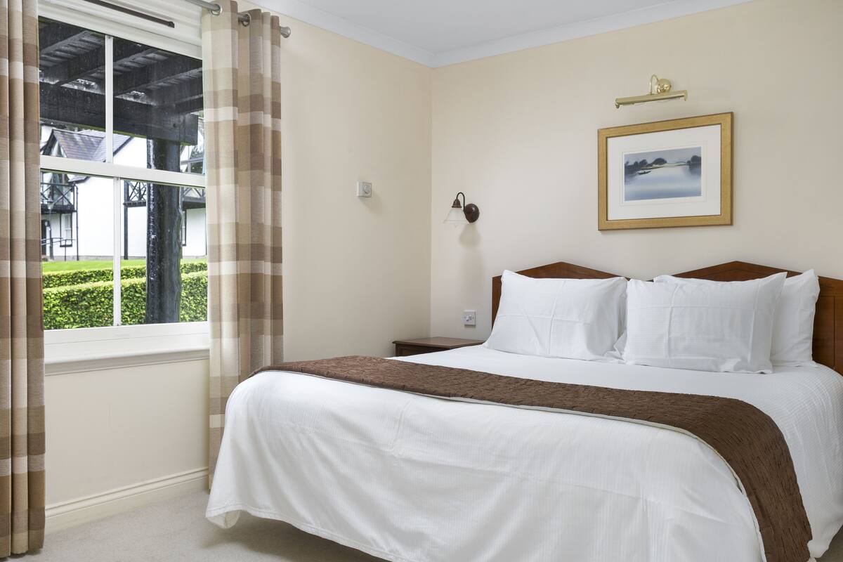 A hotel room with a bed, a window with curtains, a bedside table, a wall-mounted lamp, and a framed picture.