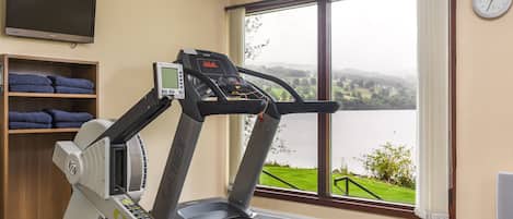 A treadmill with a digital display, a television above it, and a window with a view of a lake and greenery.
