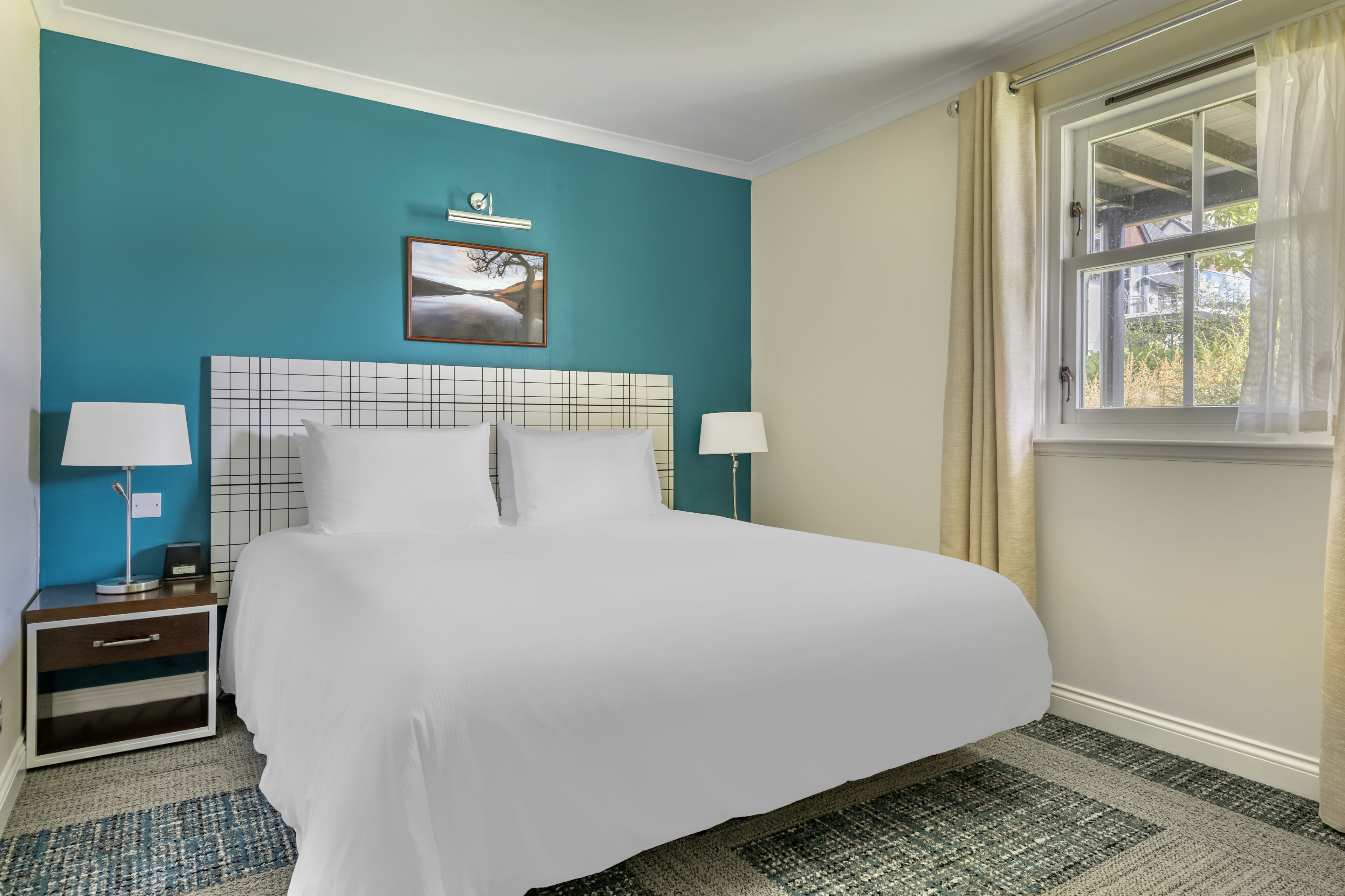 A hotel room with a large bed, two bedside tables, a nightstand, a painting on the wall, and a window with curtains.